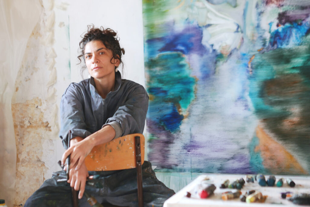 Portrait de Sarah Jérôme artiste peintre, assise sur une chaise devant une toile.